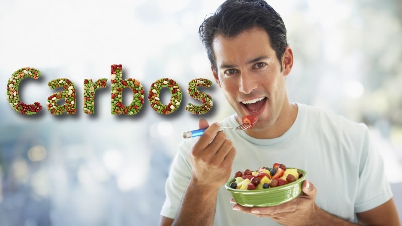 Hombre come un plato de fruta con la palabra Carbos escrita con legumbres para simbolizar las fuentes principales de carbohidratos.